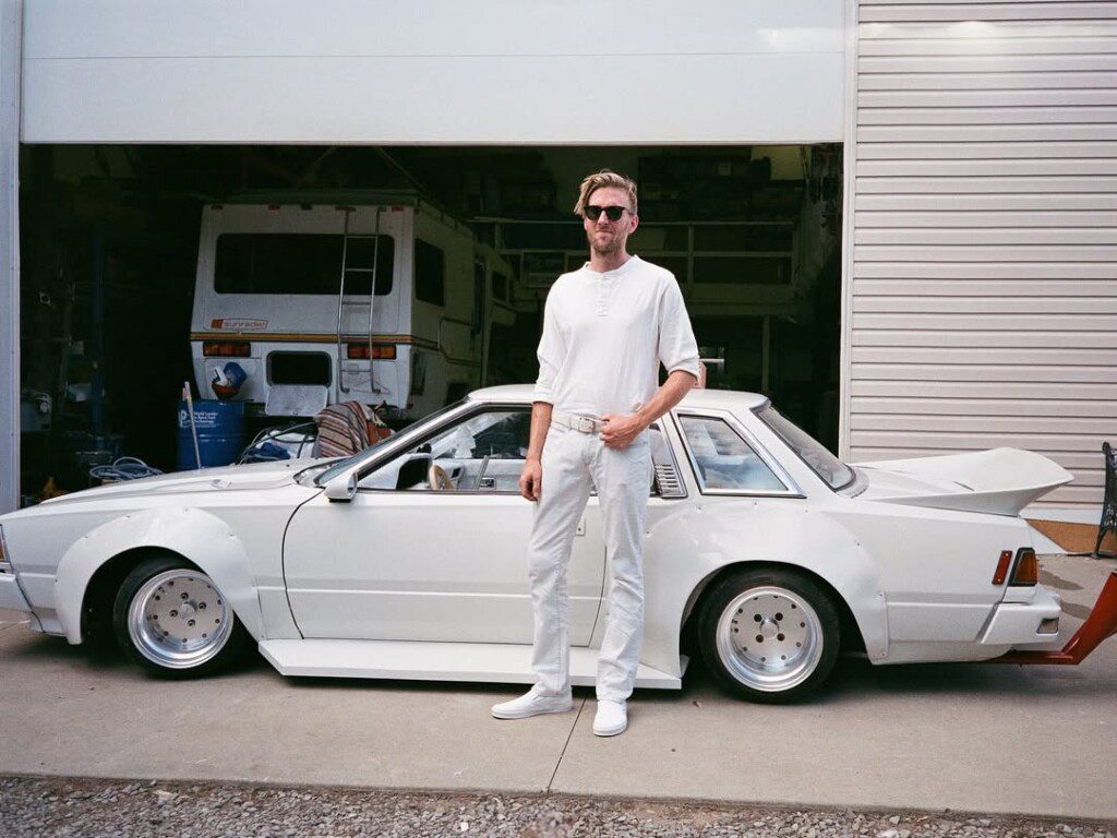 Man in white outfit stands by a modified white sports car outside a garage, exuding a stylish, automotive vibe.