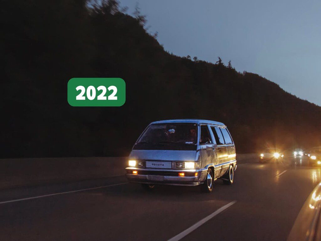 Vintage van driving on a highway at dusk with headlights on, marked 2022 in green.