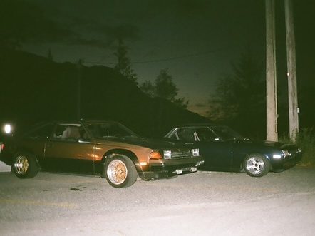 Two vintage cars parked on a dimly lit road at night, with a backdrop of trees and a dark sky.