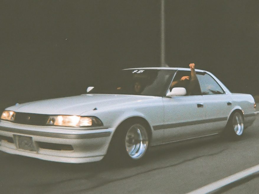 Vintage white sedan driving with arm out the window at night on a highway, showcasing classic car culture.