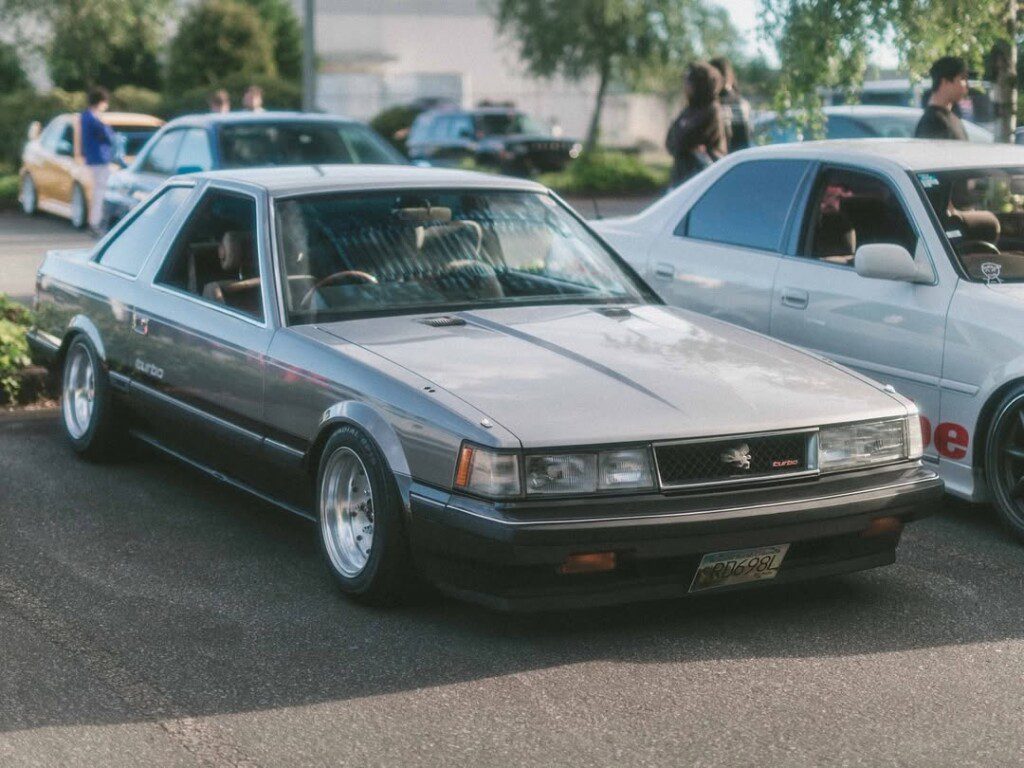 Silver vintage car at a car meet, parked next to a white sedan, with people socializing in the background.