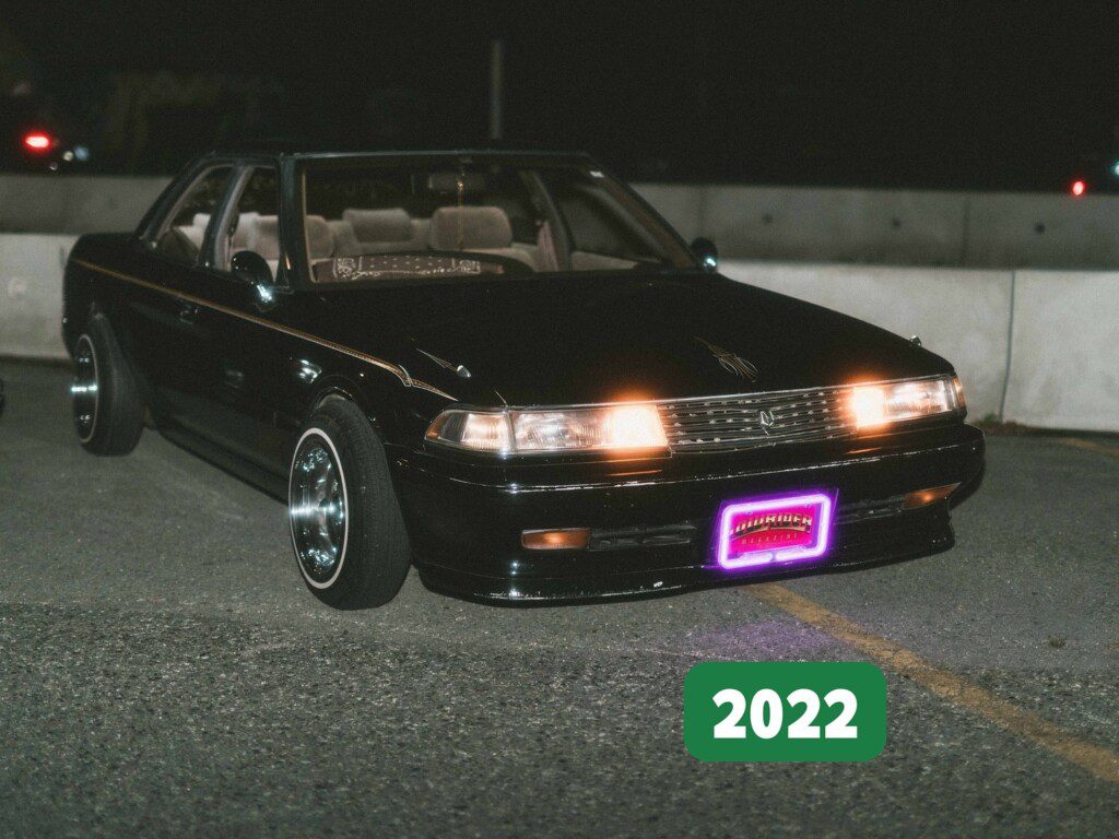 Vintage black car with vibrant headlights, parked at night, featuring a neon license plate in 2022.