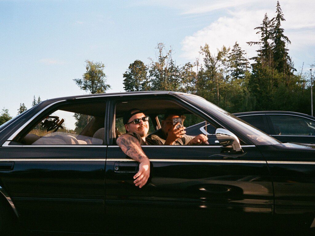 Two people in a black car, one leaning out with sunglasses and tattoos, enjoying a sunny day drive.