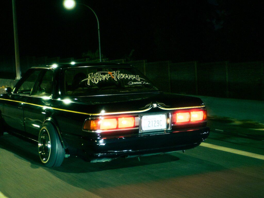 Dark car cruising at night with a glowing streetlight backdrop and stylish decals on rear window.