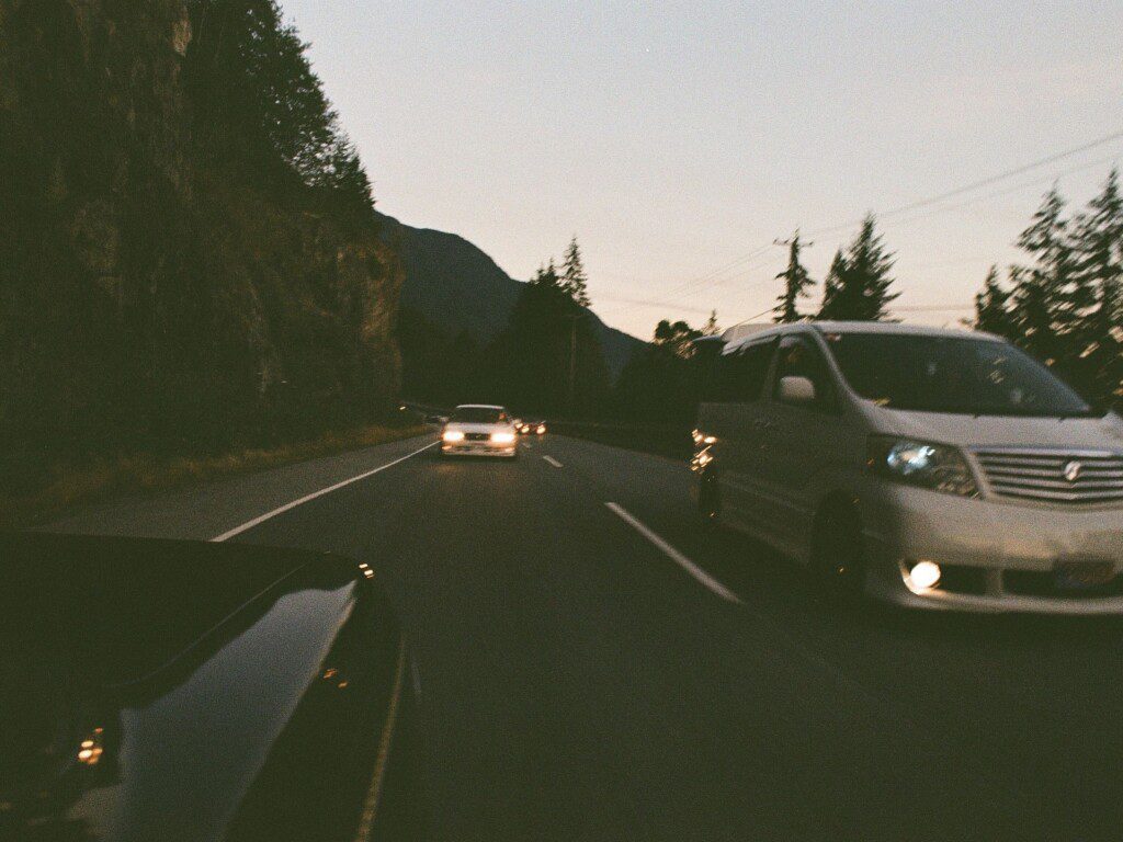 Cars driving on a winding mountain road at dusk, surrounded by trees and rocky cliffs.