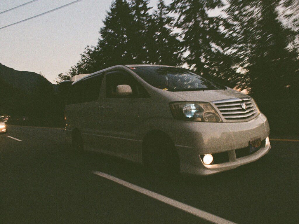 White van driving on a tree-lined road at dusk, headlights on, showcasing smooth travel in serene, natural setting.