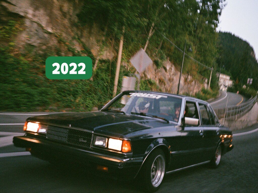 Vintage car driving on a scenic road in 2022, surrounded by lush greenery and mountains.
