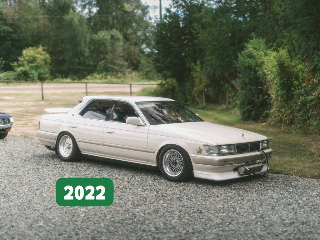 Classic white sedan parked on gravel driveway with greenery, 2022. Vintage car show setting.