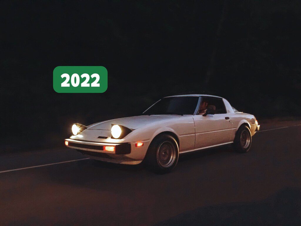 White classic sports car with pop-up headlights driving at night, 2022 green sign in background.