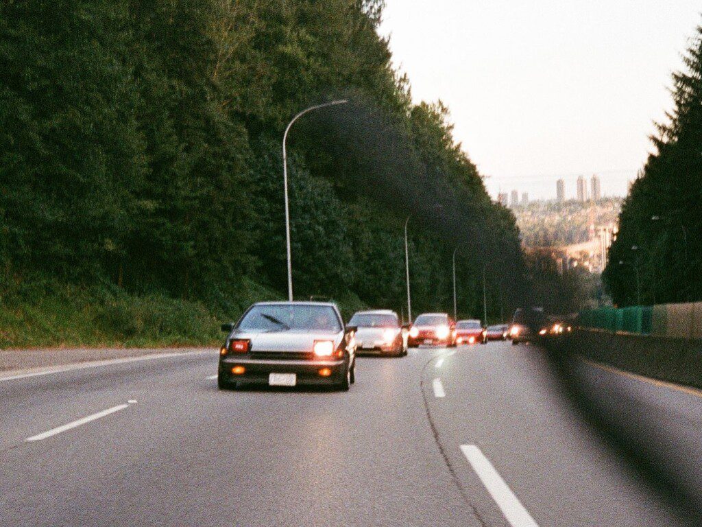 Cars driving on a forest-lined highway at dusk, with city skyline in the background.
