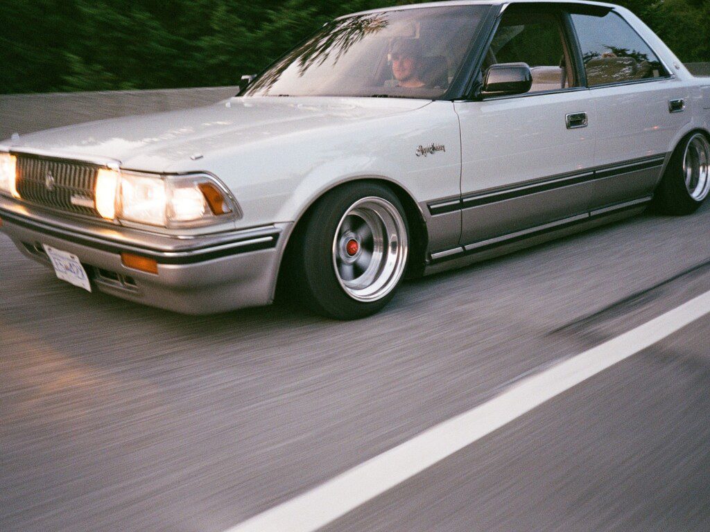 Vintage white sedan cruising on the highway, featuring lowered suspension and custom wheels.