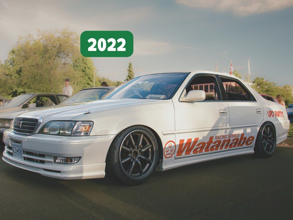 Customized white sedan at a car meet, featuring Watanabe decals and modified wheels, with 2022 in the background.