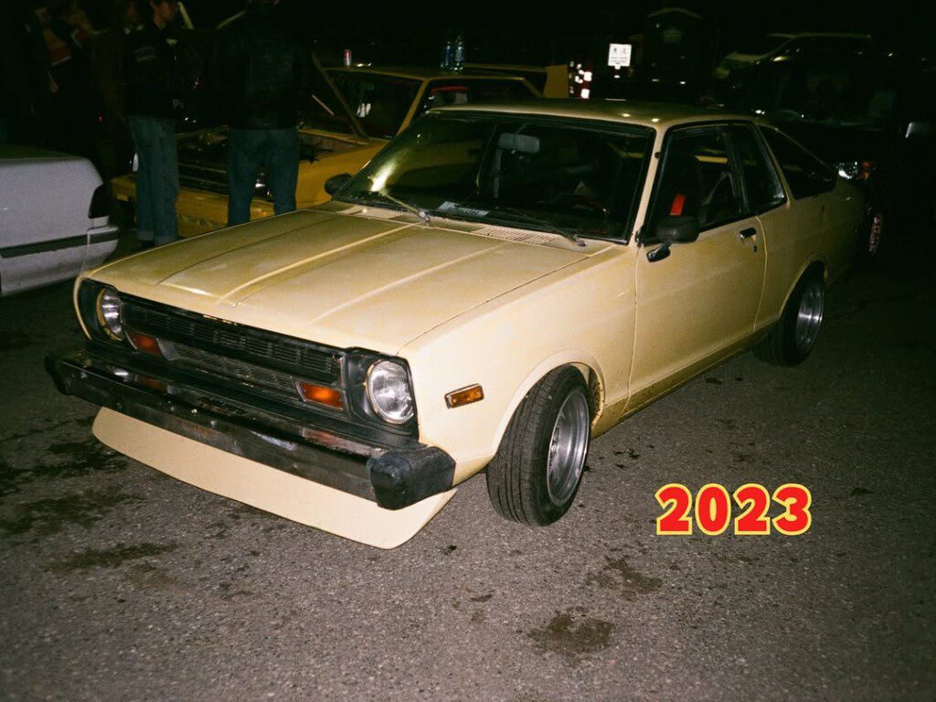 Classic yellow car in a nighttime gathering, parked among others, with bold 2023 in the foreground.
