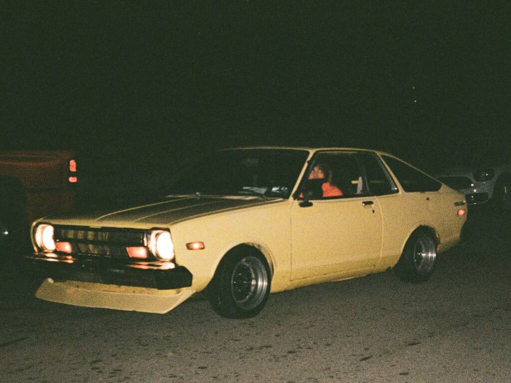 Vintage yellow car driving at night with headlights on, set against a dark background.
