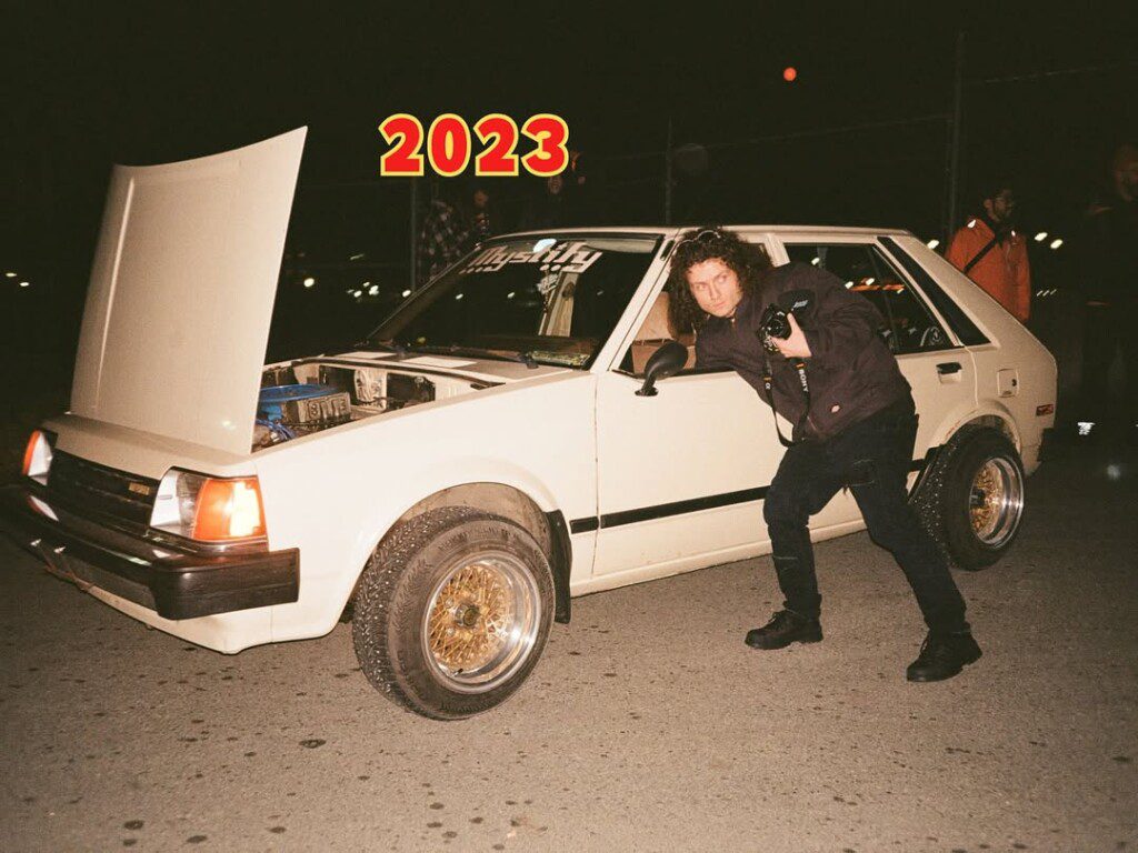 Vintage car with open hood at night, posing photographer, vibrant 2023 text; urban automotive scene.