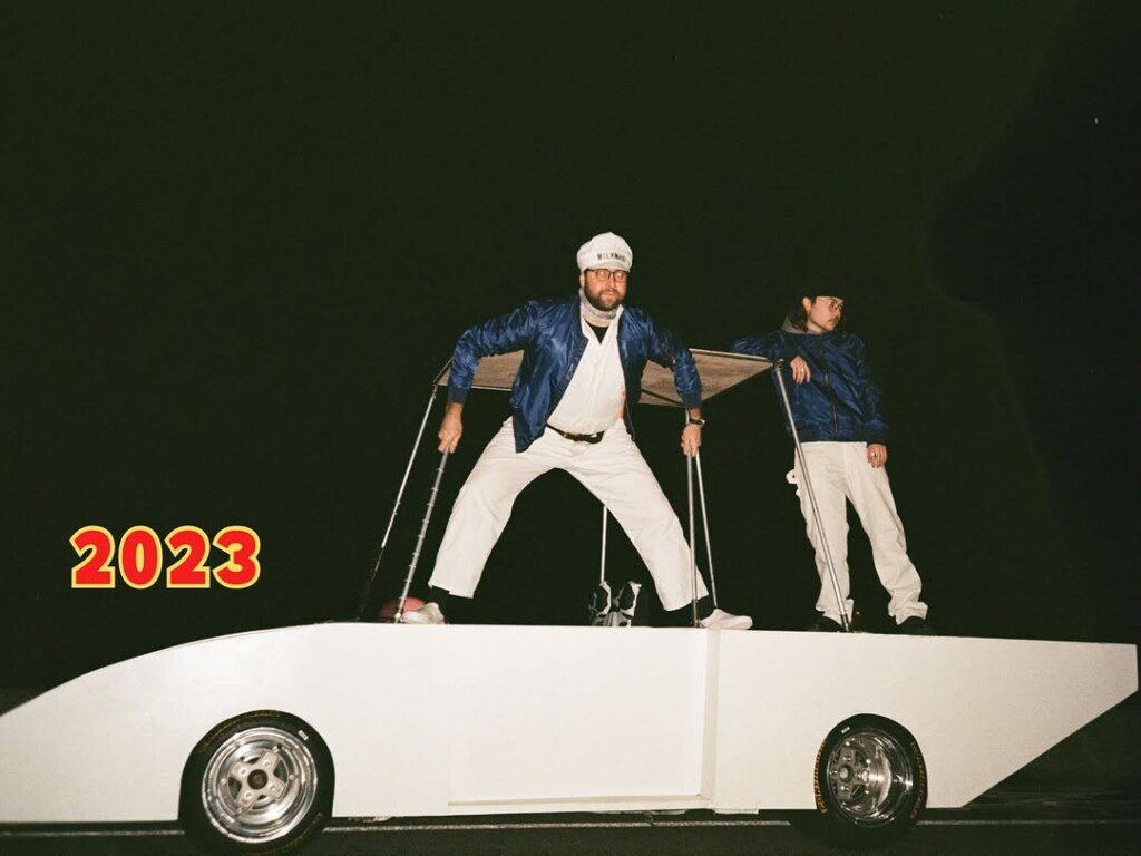 Two people in blue jackets stand on a futuristic car against a dark background. Text reads 2023.