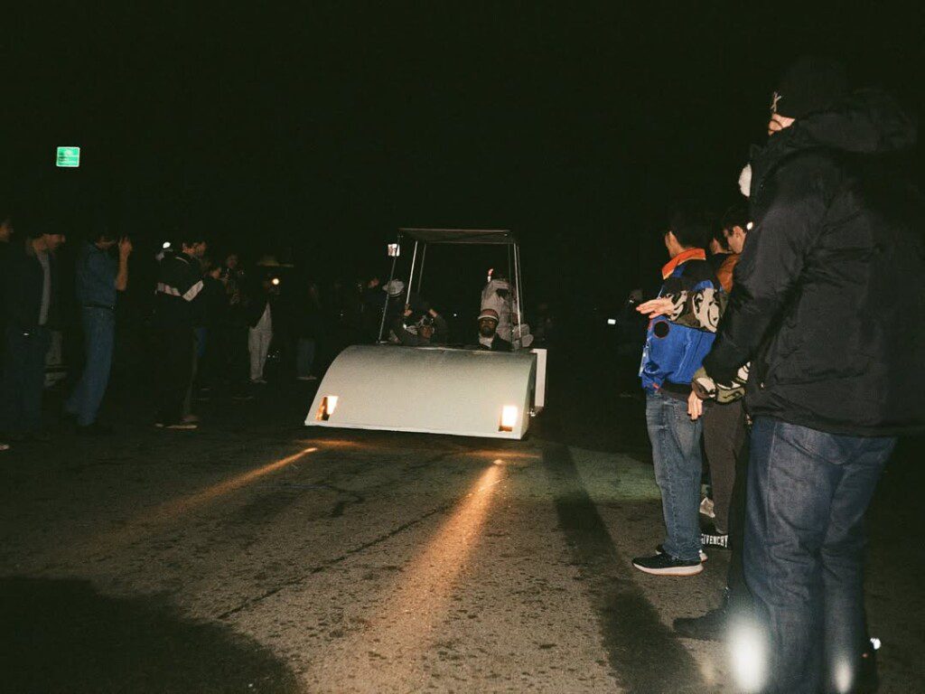 Nighttime race with a futuristic car and crowd of spectators on a dimly lit street.