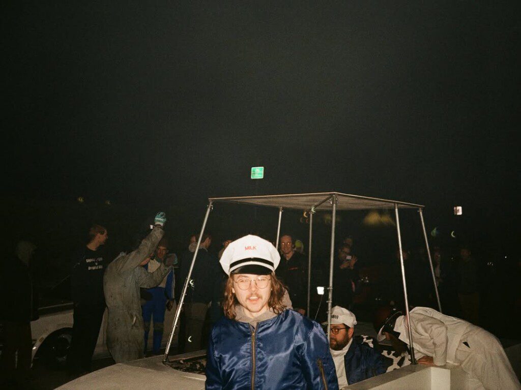 Group of people in costumes gathered around a cart at night, featuring milkman attire and festive atmosphere.