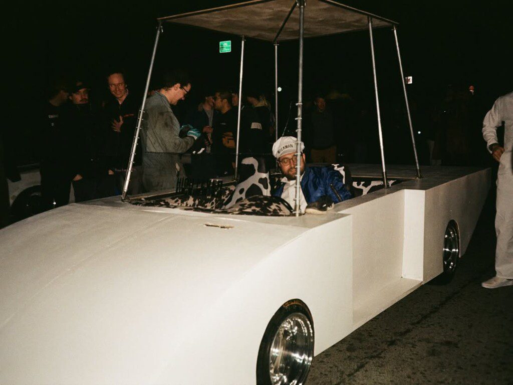 Man in creative car costume resembling Flintstones vehicle at night event, dressed in blue jacket and cap.