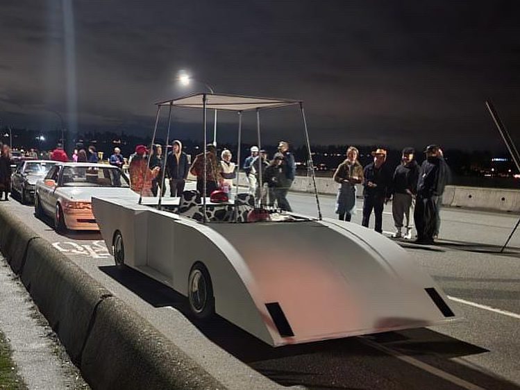 Group gathered around Flintstones car replica at night on road.