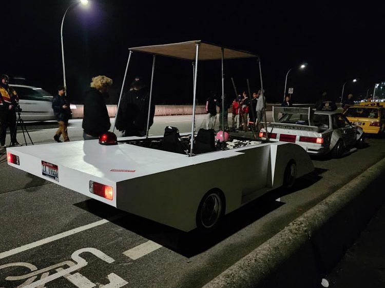 Unique custom car with minimalistic design parked on a dimly lit street at night, surrounded by people.