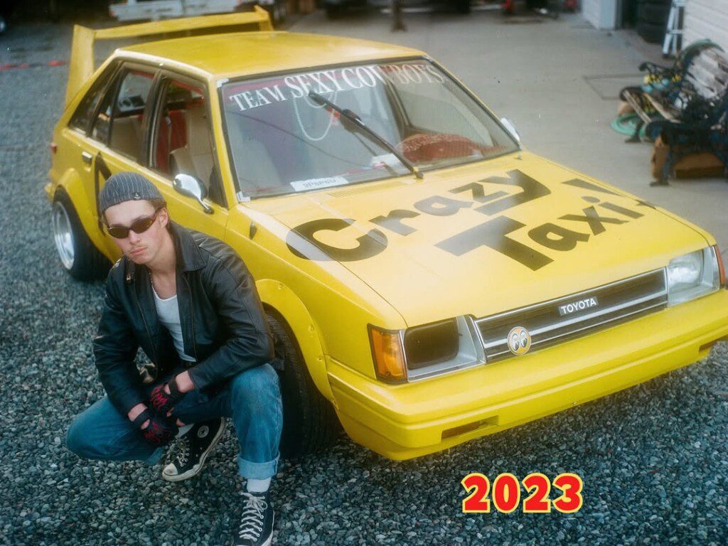Person in leather jacket poses by a yellow Toyota Crazy Taxi car with Team Sexy Cowboys sign.