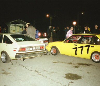 Group of people gathered around vintage cars at night, featuring a yellow car with racing number 777.