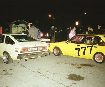 Group of people gathered around vintage cars at night, featuring a yellow car with racing number 777.