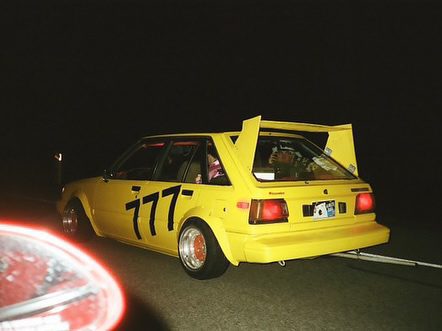 Yellow race car with 777 on the side, driving at night on a racetrack, showcasing a sporty design and large rear spoiler.