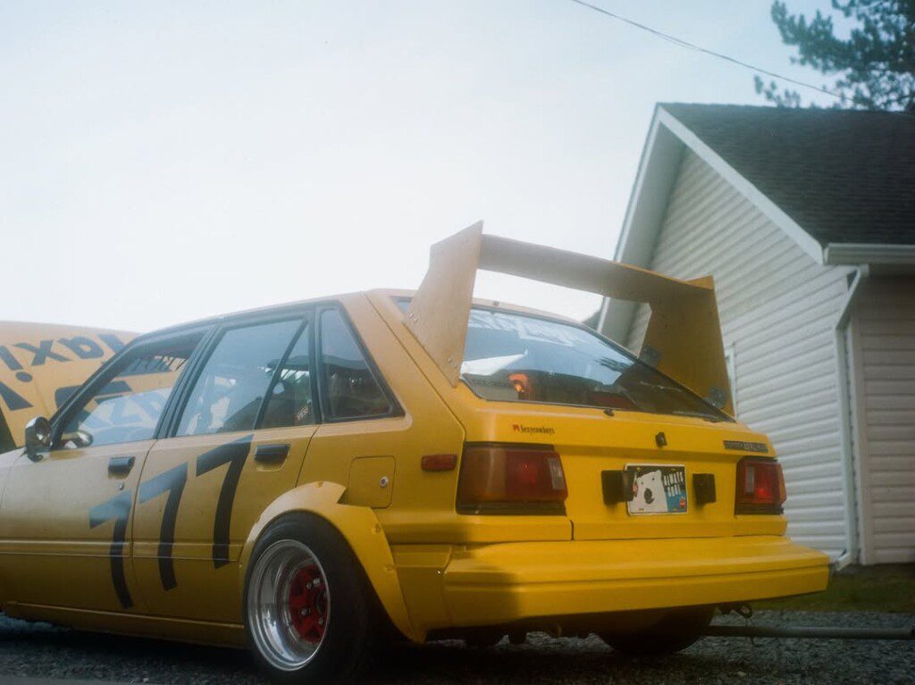 Yellow race car with number 777, large rear spoiler, parked near a house. Racing modification details visible.