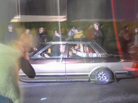 Car with passengers displaying peace signs surrounded by people at night under bright lights.