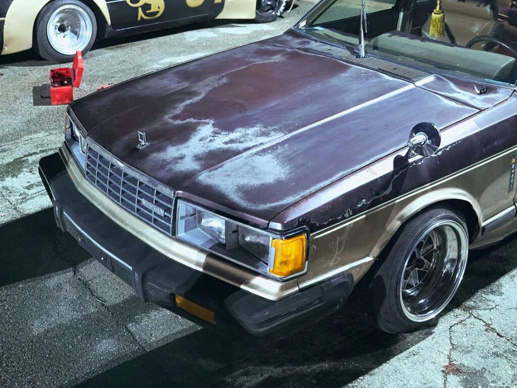 Vintage custom car with rust and unique paintwork at a nighttime meetup, showcasing retro automotive style.