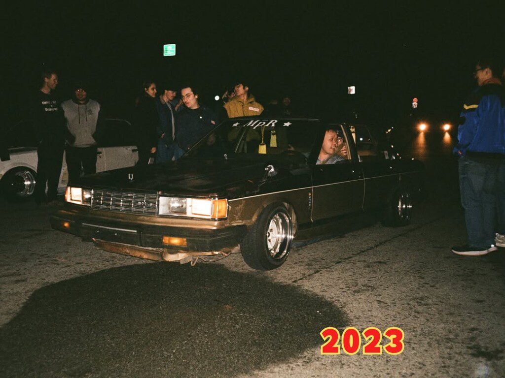 Vintage car meetup at night with people gathered, featuring a classic sedan, 2023.