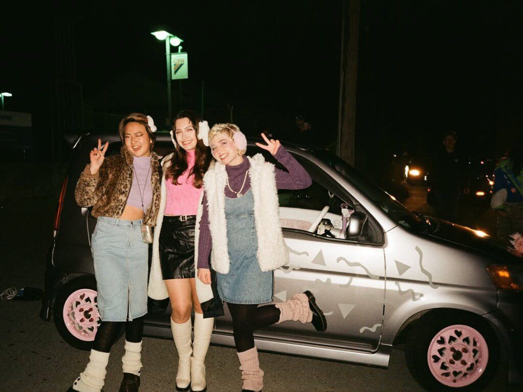 Three friends pose in stylish outfits near a decorated car at night, expressing joy and friendship with peace signs.
