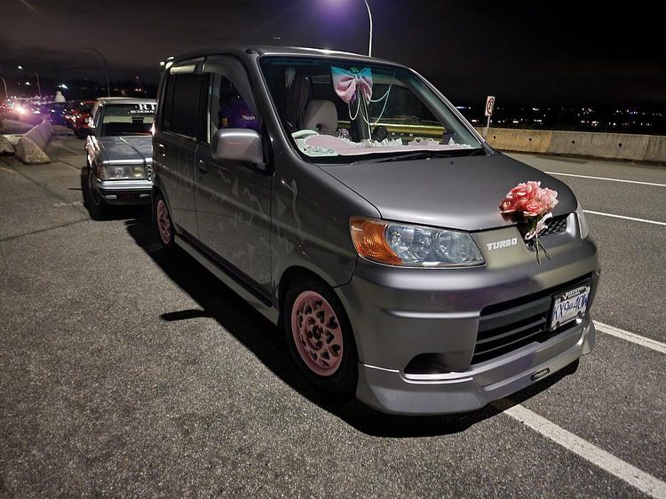 Decorated grey minivan with pink rims and flowers parked on a street at night.