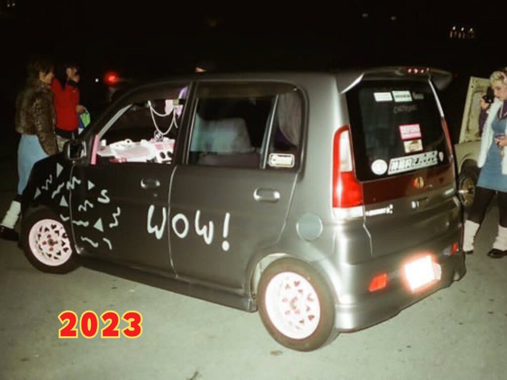 Modified car with Wow! painted on the side, surrounded by people at night, 2023 style.