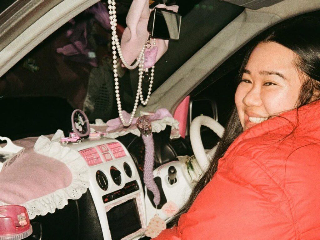 Smiling person in a car with a pink and cute, customized interior adorned with accessories and plush decorations.