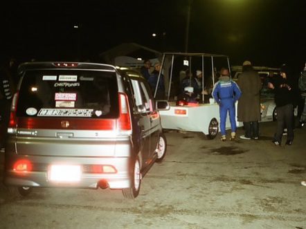 Group of people at a nighttime car meetup, featuring a modified vehicle and parked cars in the background.