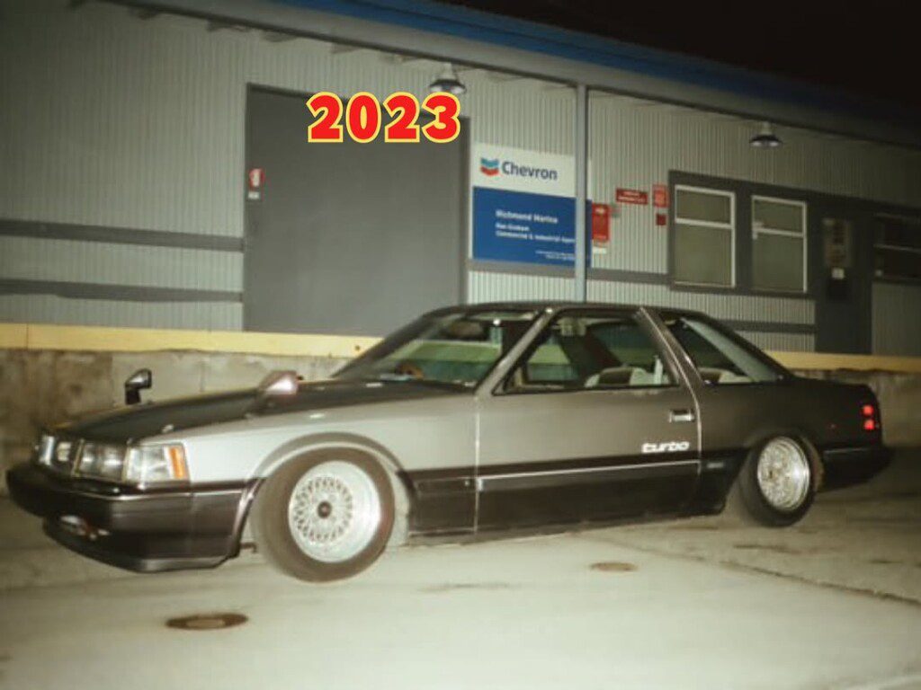 Classic silver turbo car parked at night near industrial building with 2023 text overlay.