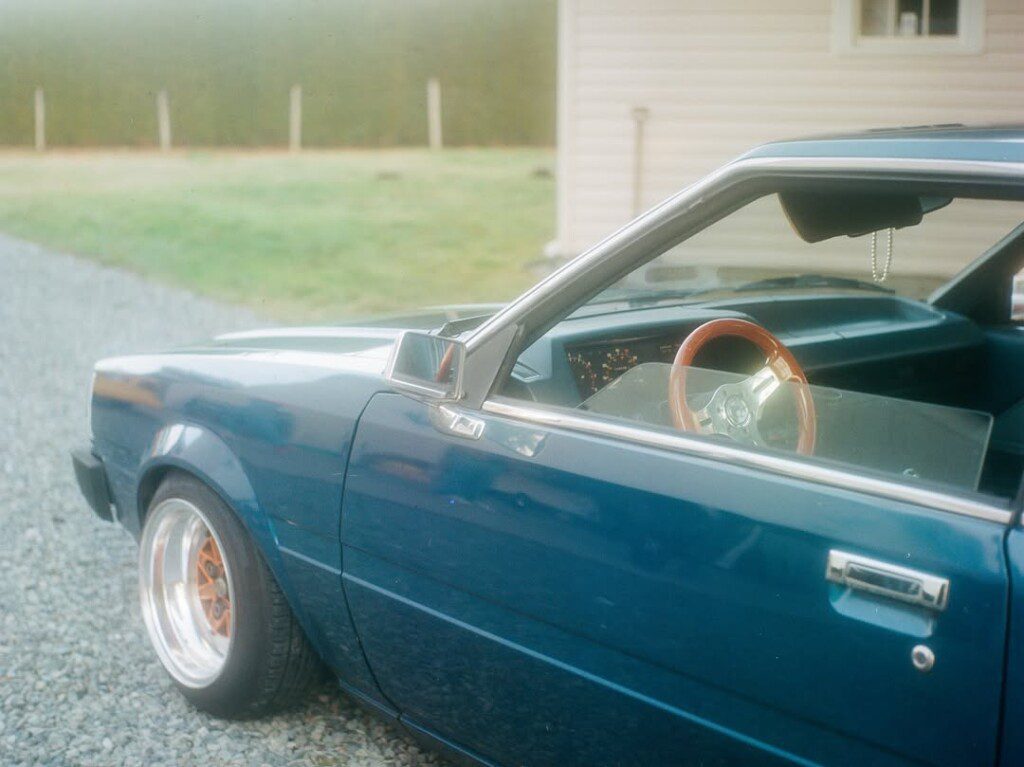 Classic blue car with open door and wooden steering wheel on gravel driveway. Vintage automotive style.