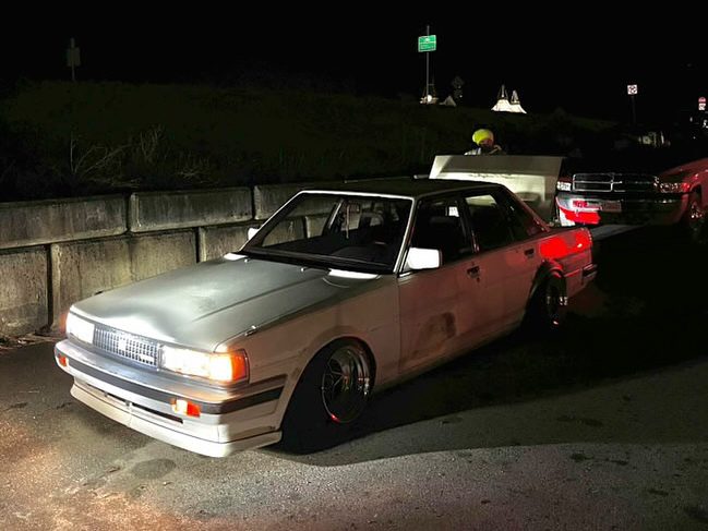 Vintage white sedan parked on roadside at night, illuminated by streetlights, with police vehicles in the background.