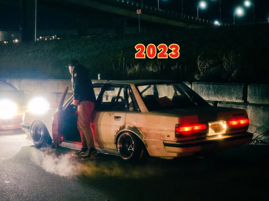 Late-night car scene with glowing headlights and a person exiting a vehicle, text 2023 above. Urban ambiance.