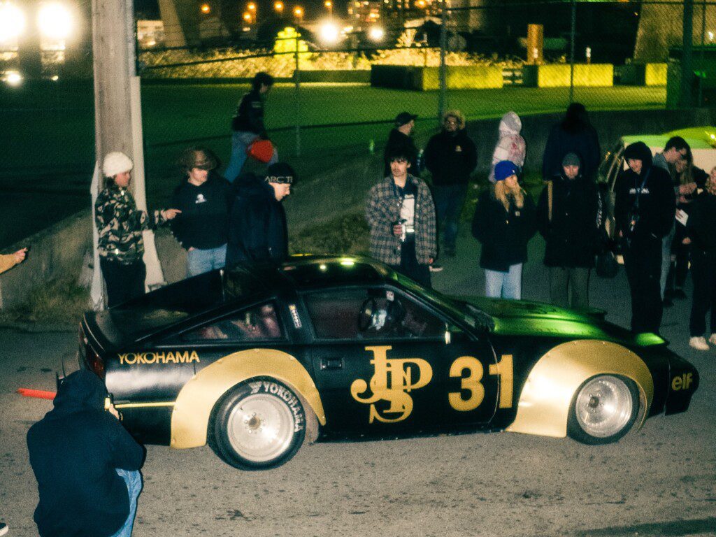 Vintage JPS 31 race car at night event, surrounded by onlookers in winter attire, under bright streetlights.