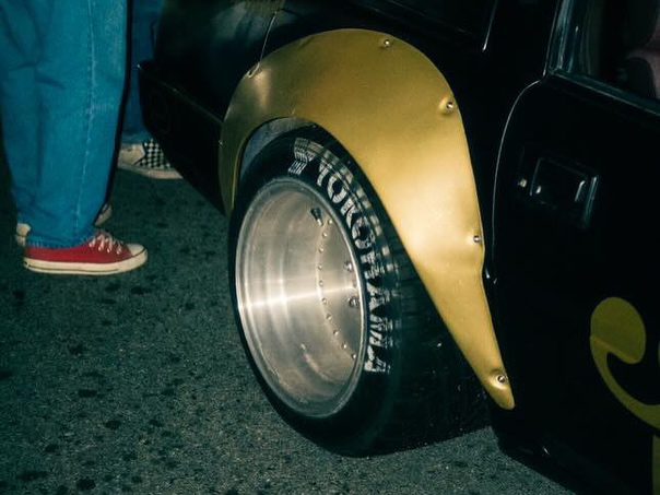 Close-up of a modified car's back wheel and fender with nearby people wearing casual shoes.