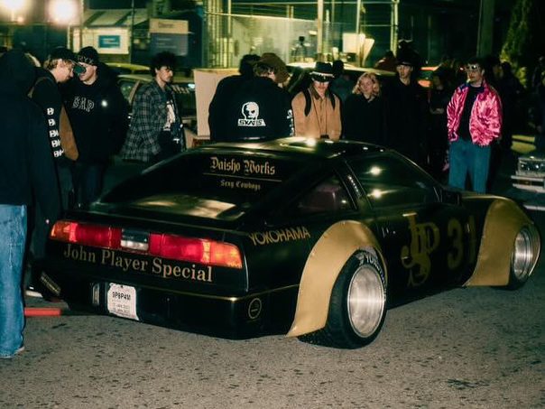 Custom black and gold sports car at a night car meet, surrounded by people, featuring John Player Special branding.