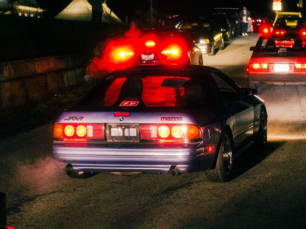 Night scene of a blue Mazda RX-7 car on a road, emitting exhaust vapor, with glowing taillights.