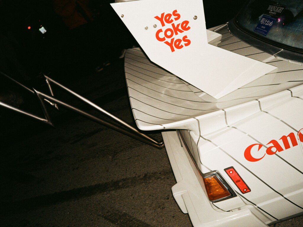 Vintage white car with Yes Coke Yes decal, striped design, and large rear spoiler, parked at night.