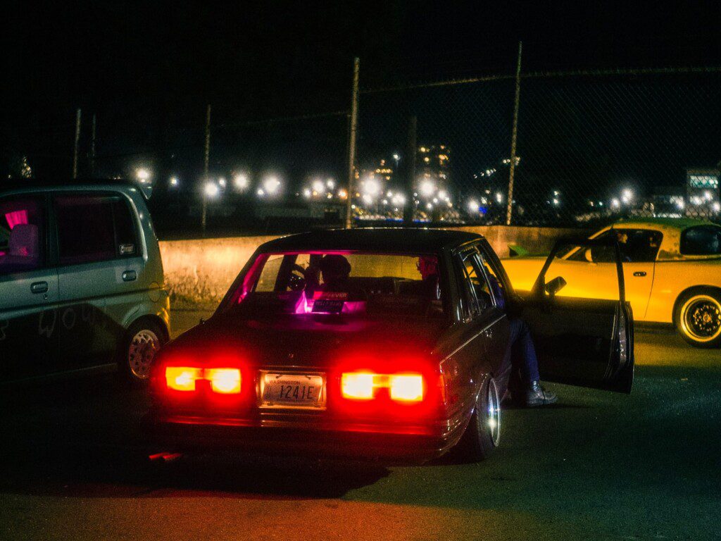 Vintage cars with bright taillights and open doors in a nighttime urban setting.