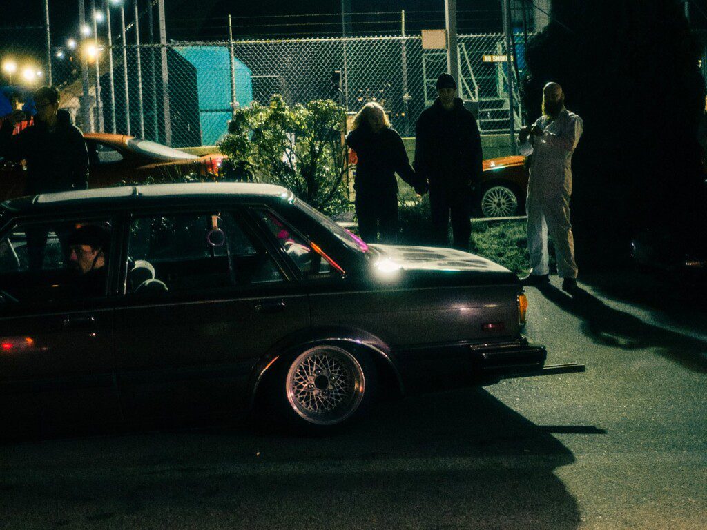 Vintage car at night with silhouettes of people and a brightly lit background.