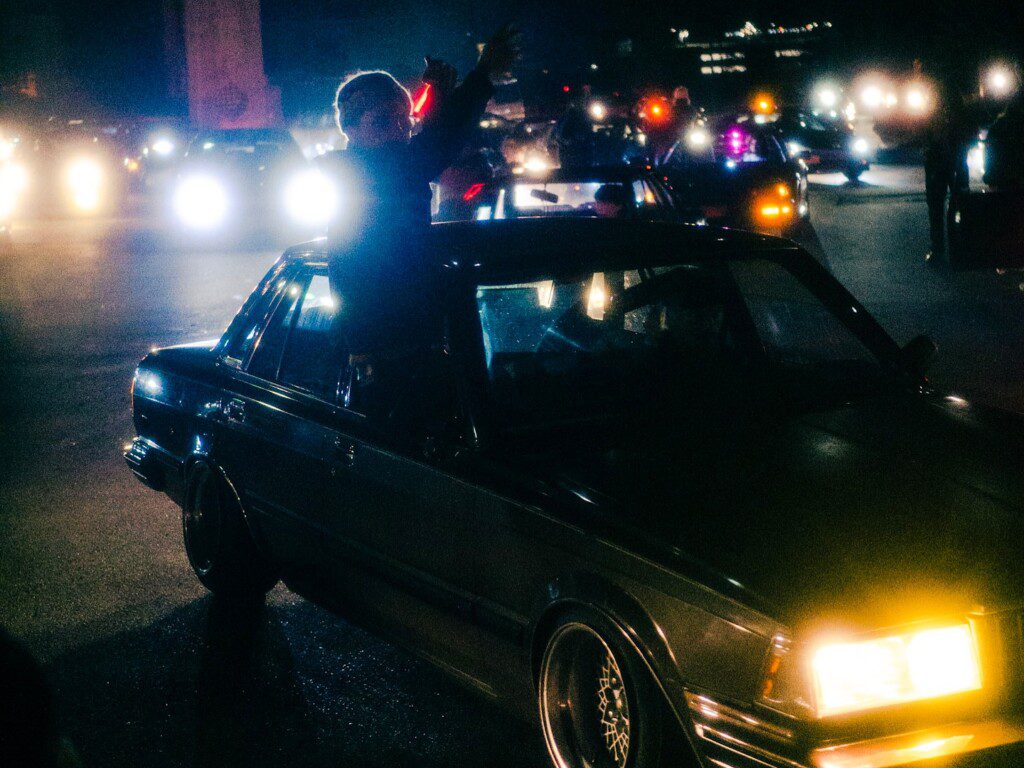 Car with people celebrating on a busy night street, headlights illuminating the scene, creating a vibrant nightlife atmosphere.
